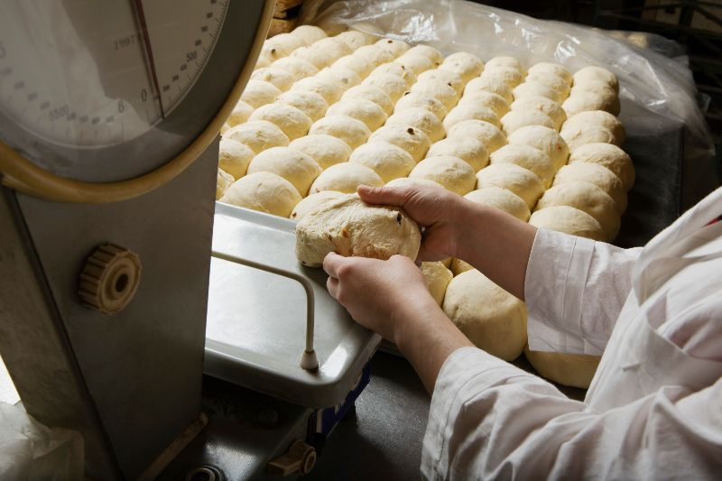 A factory worker weighing a ball of dough on a scale with rows of dough pieces in the background.