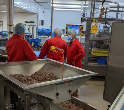 Three workers in PPE monitor industrial chocolate processing equipment in a manufacturing facility.