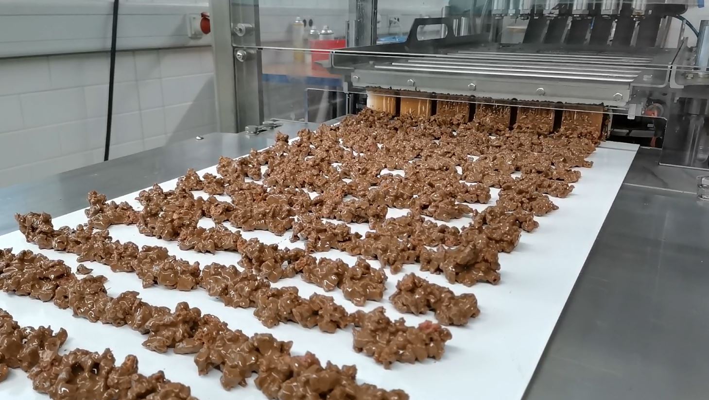 Chocolate clusters coated in melted chocolate on a conveyer belt in a factory setting.