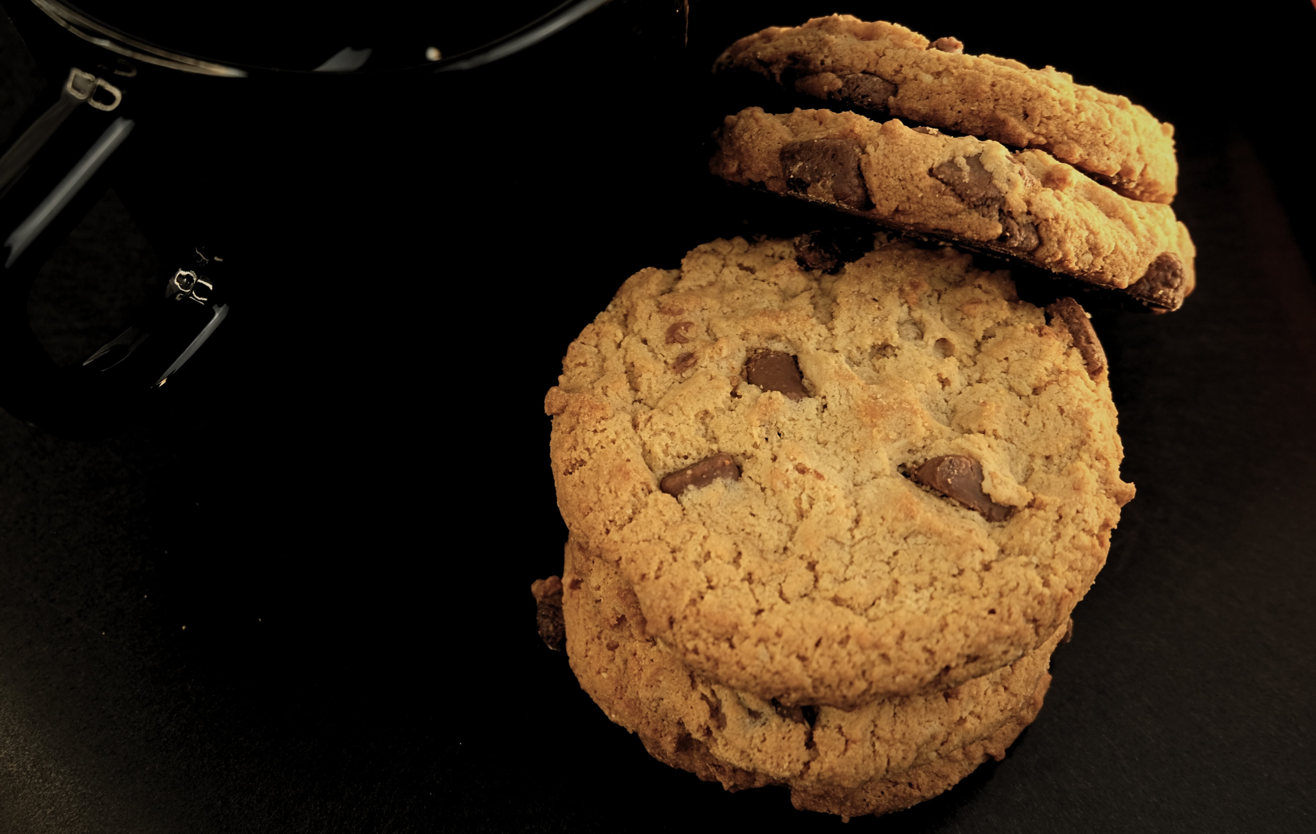 Four chocolate chip cookies next to a black cup of coffee, on a dark background.
