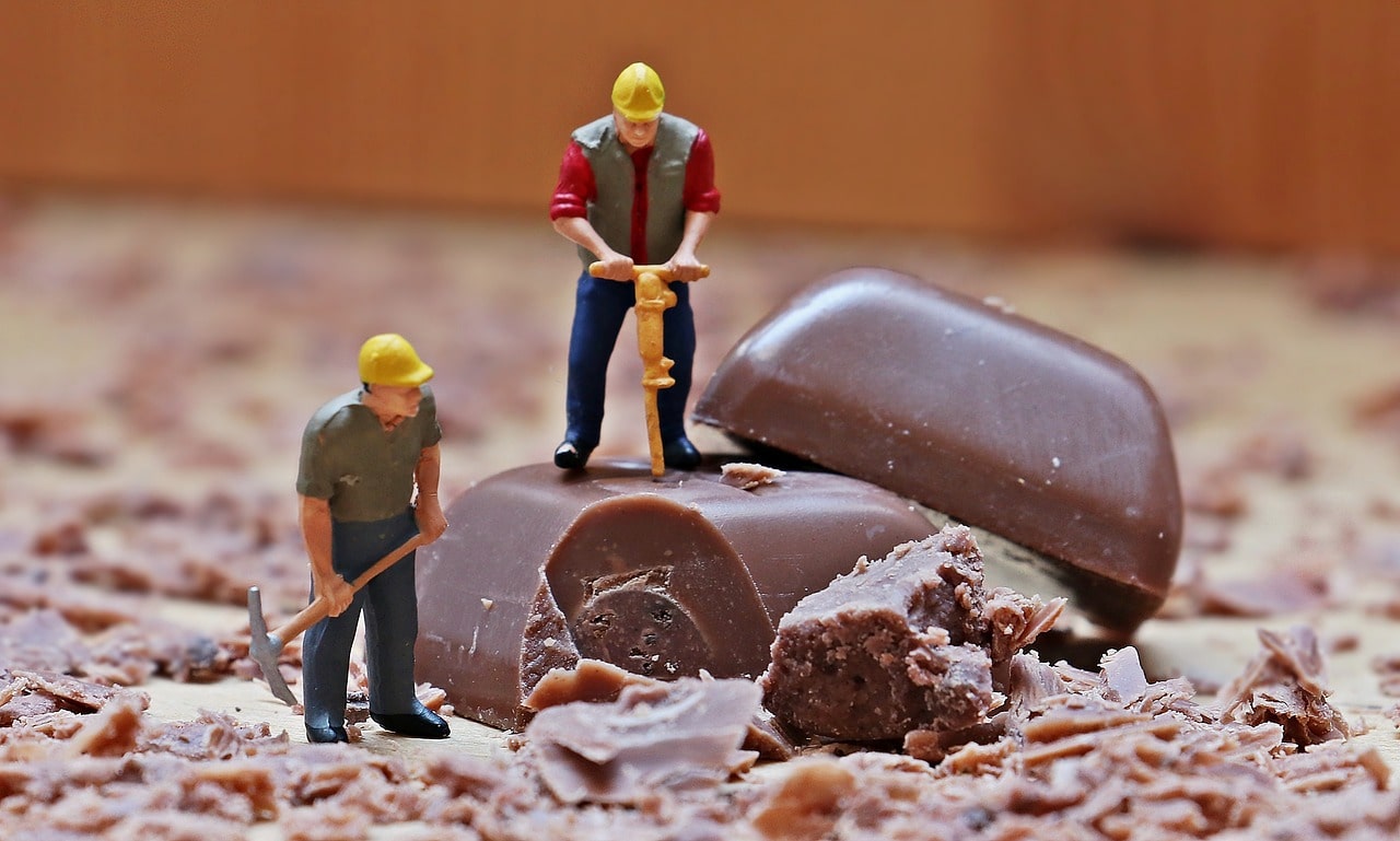 Miniature toy construction workers breaking apart chocolate pieces surrounded by shavings.
