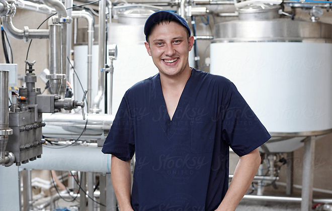 A male factory worker stands in front of industrial food processing machinery smiling.