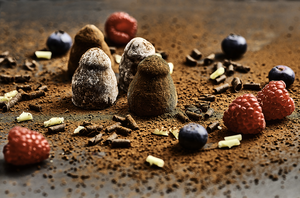 Four chocolate truffles dusted with milk and white toppings, placed on a bed of chocolate powder.