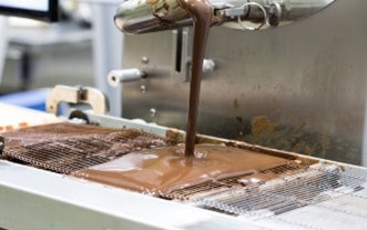 Melted chocolate flowing from processing equipment onto a wire mesh conveyor.