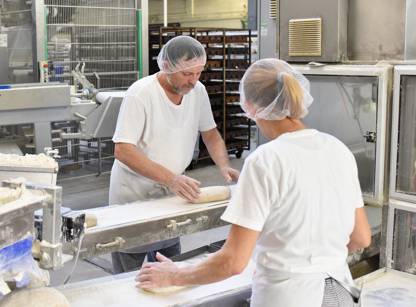 Two bakery workers shaping loaves of dough on a production line in an industrial bakery.