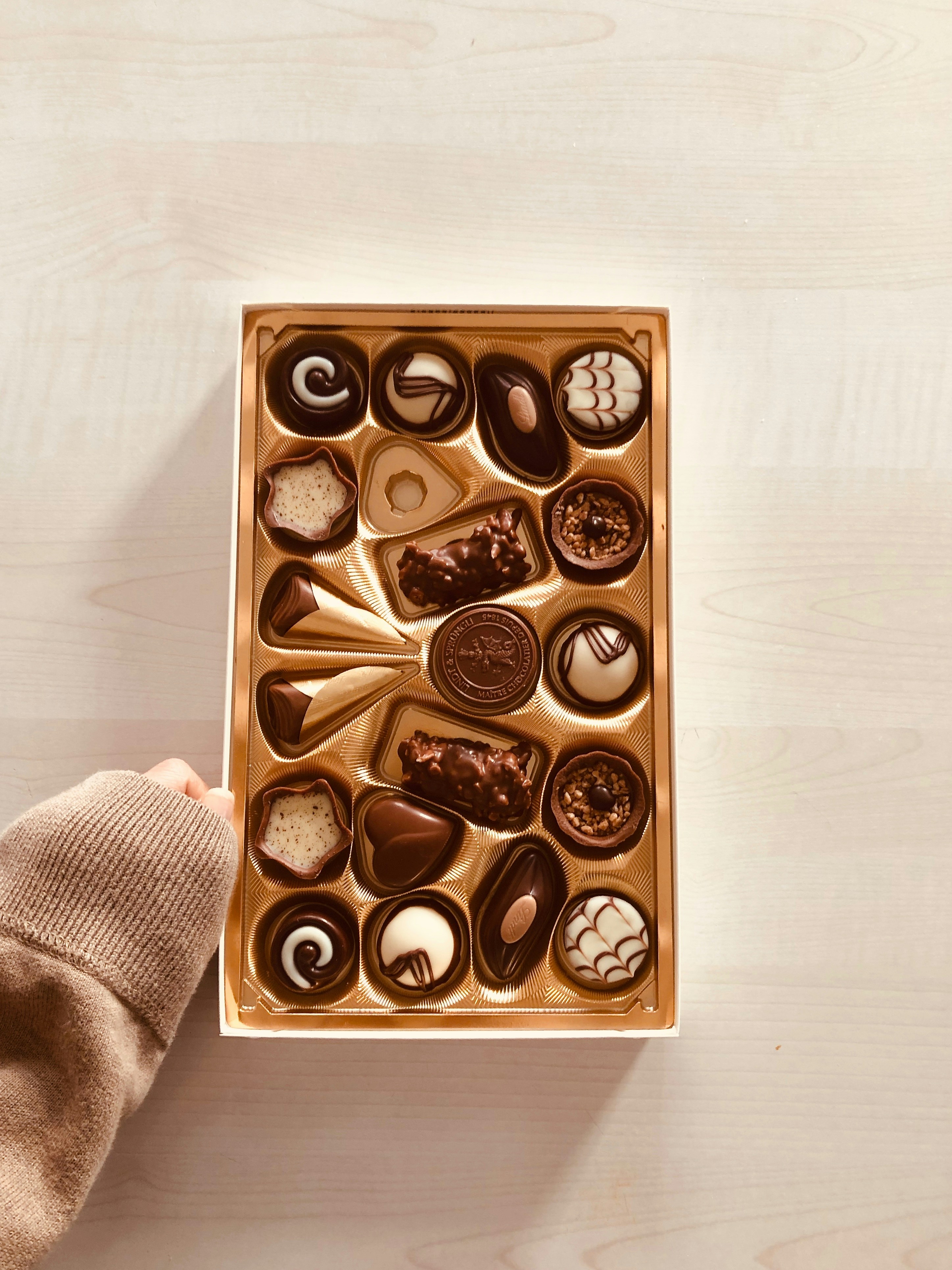 Assorted chocolates in a gold foil-lined box on a wooden surface. One is missing.