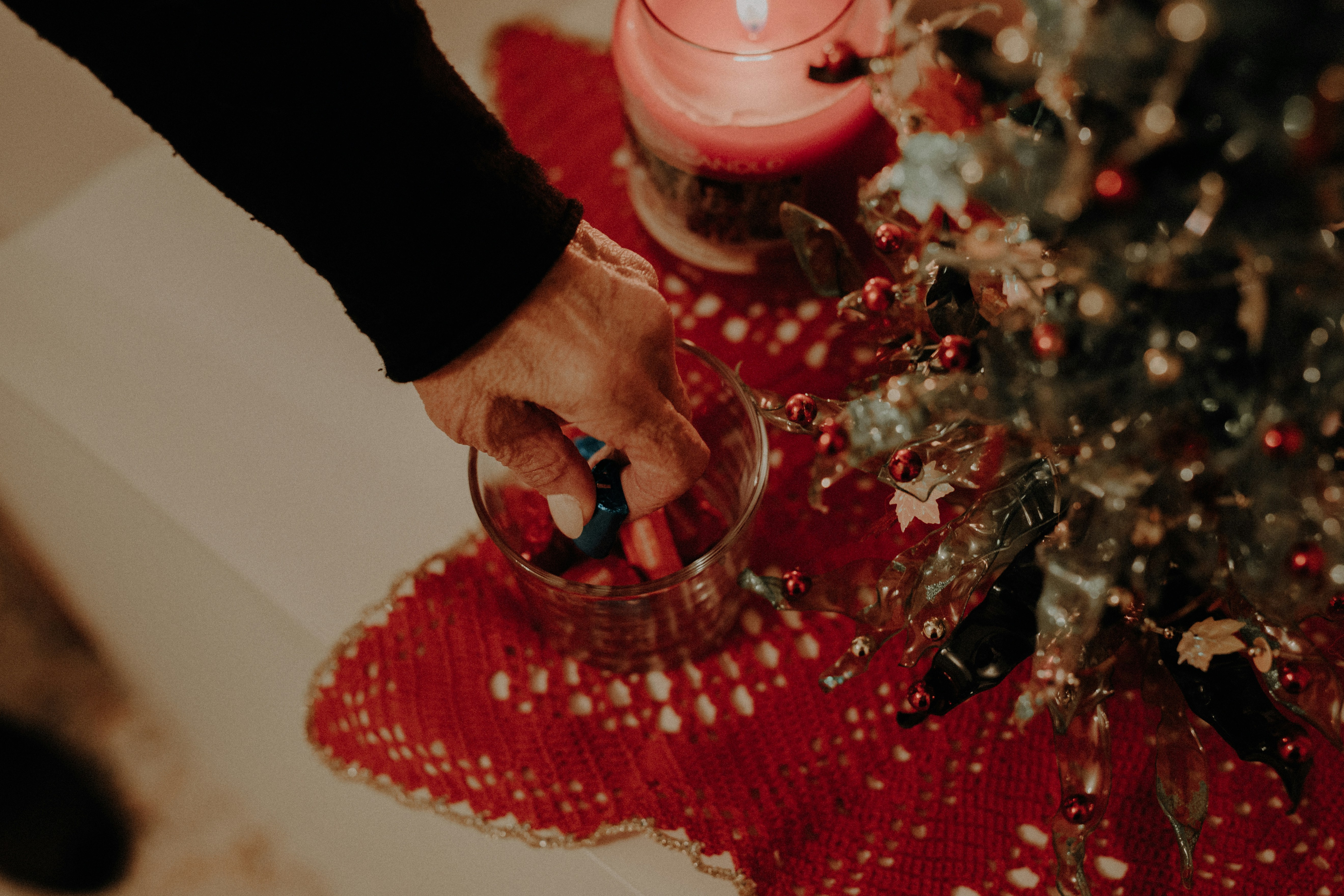 Hand reaching into a glass bowl of wrapped chocolates on a table with a Christmas tree and candle.
