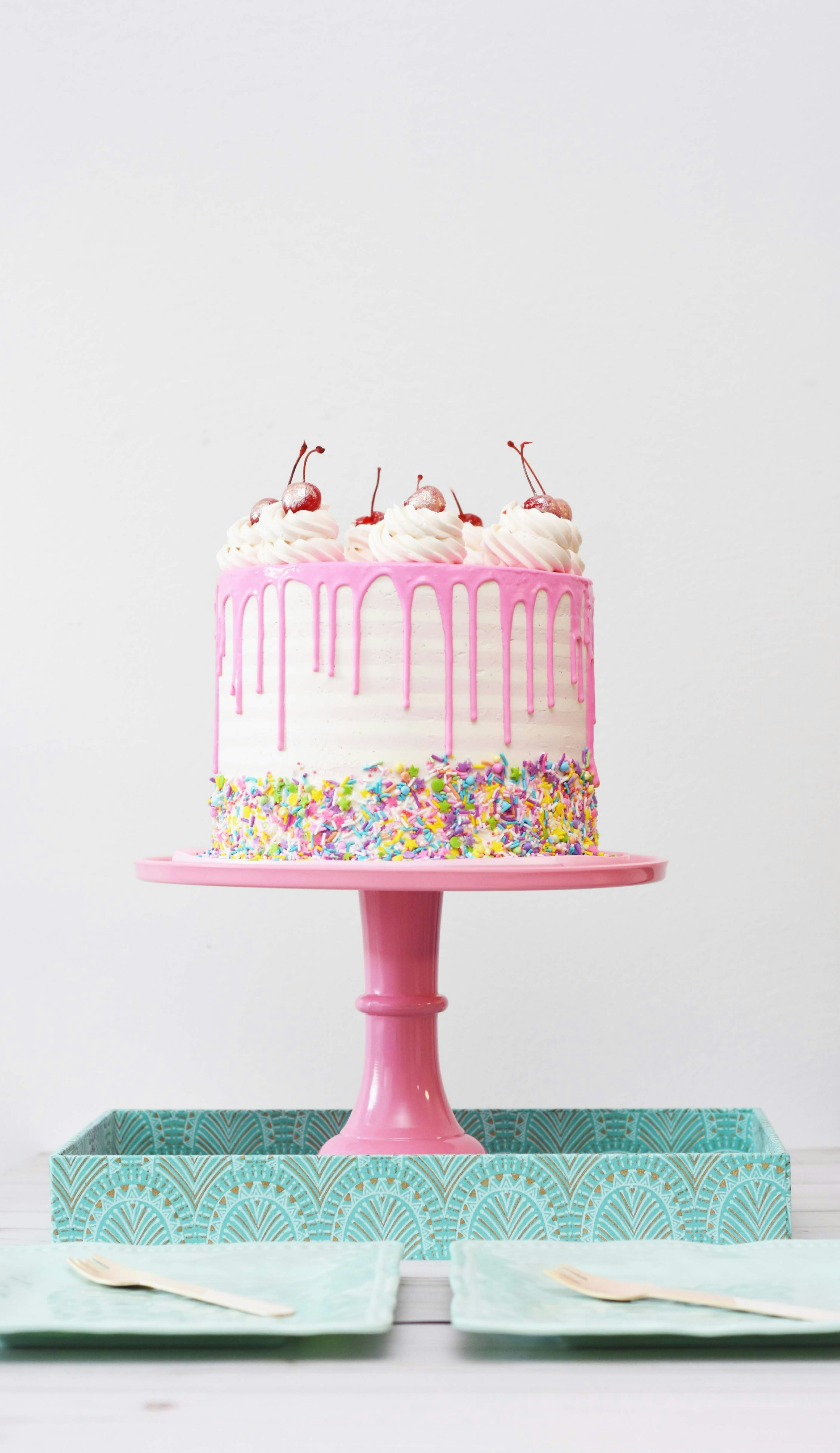 Celebration cake with pink dripping icing, sprinkles, and whole cherries on a cake stand.