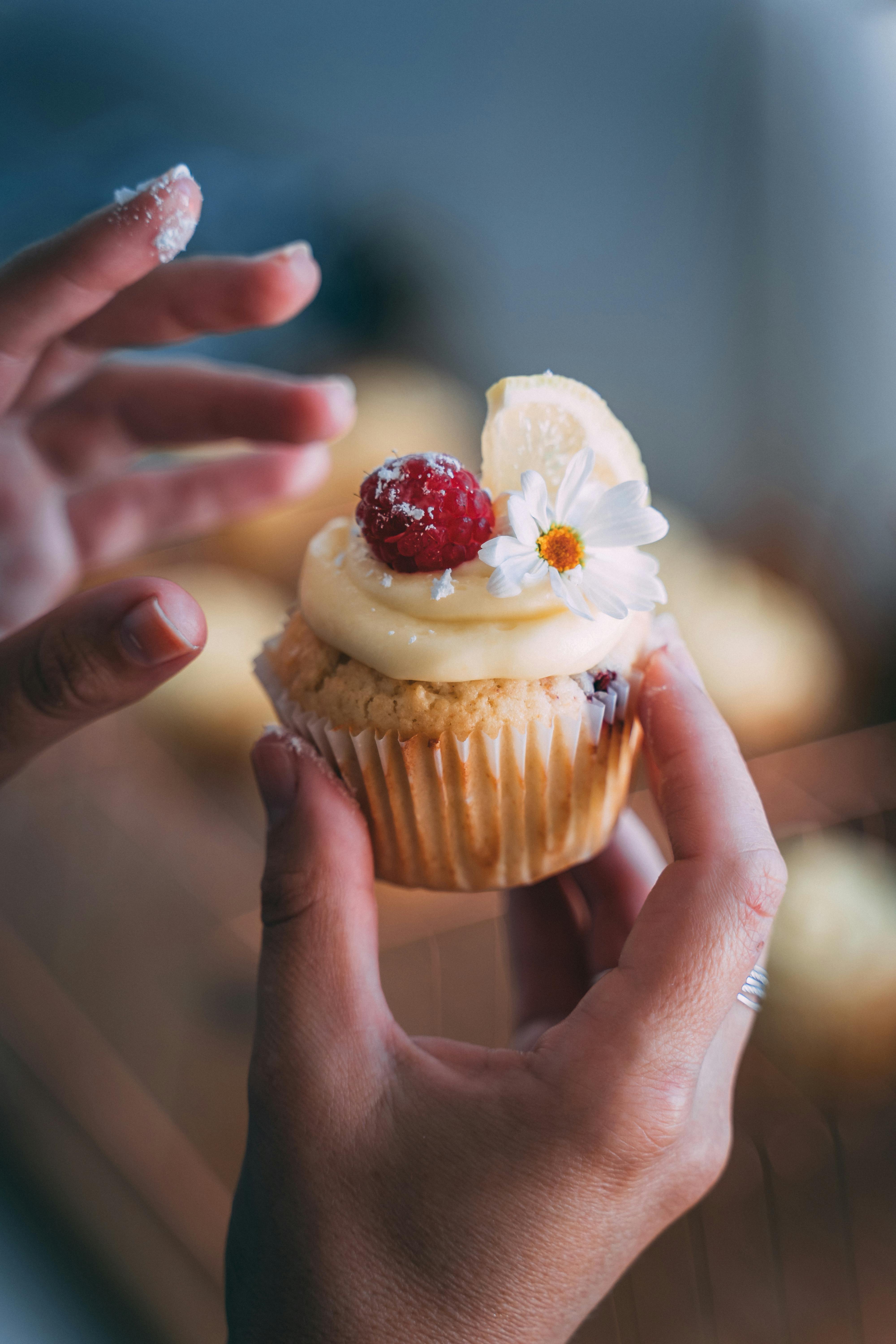 Hands holding a cupcake topped with piped icing, a raspberry, and an edible flower.