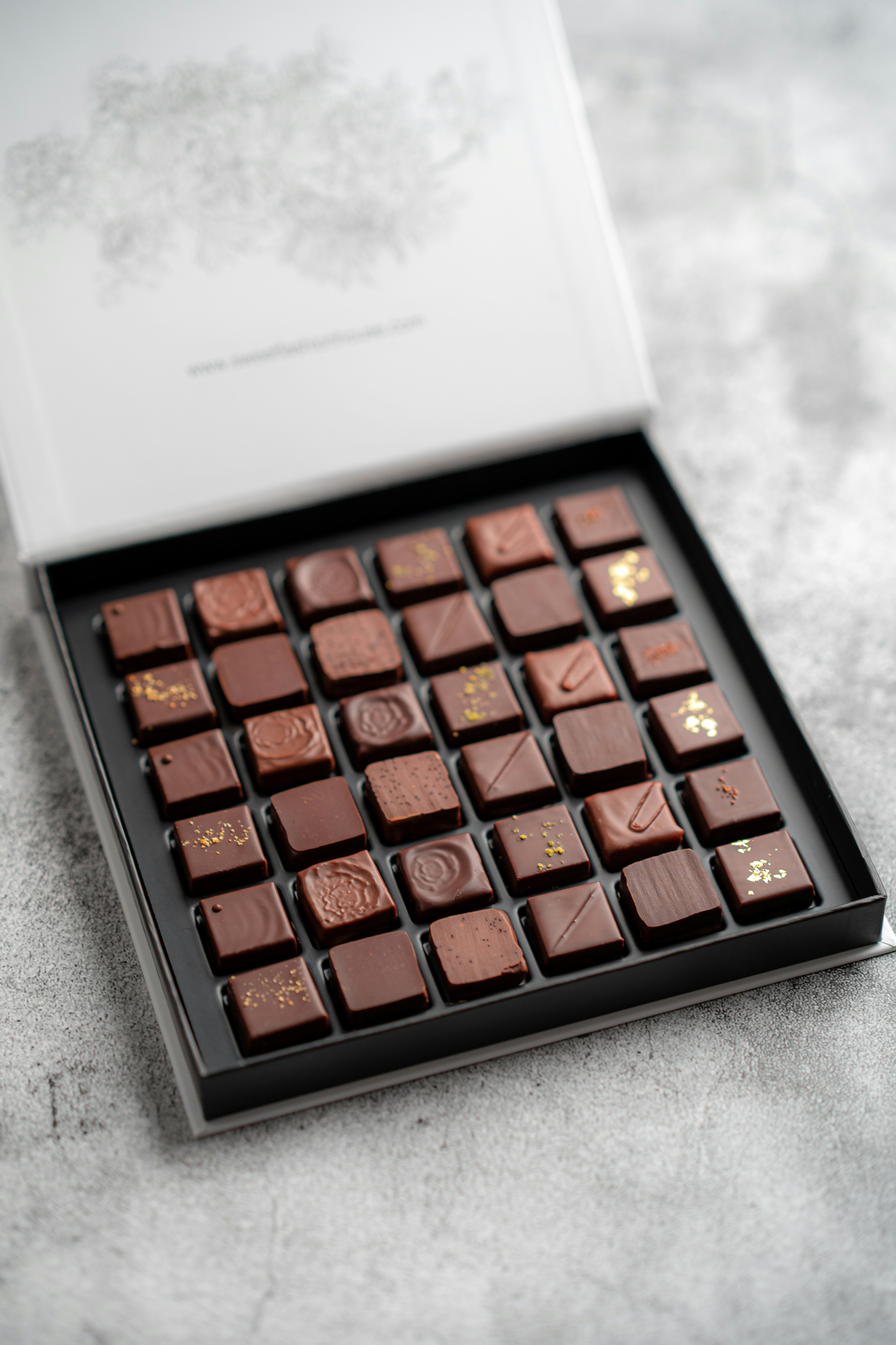 Large open box of square-shaped chocolates with assorted decorations on a light surface.