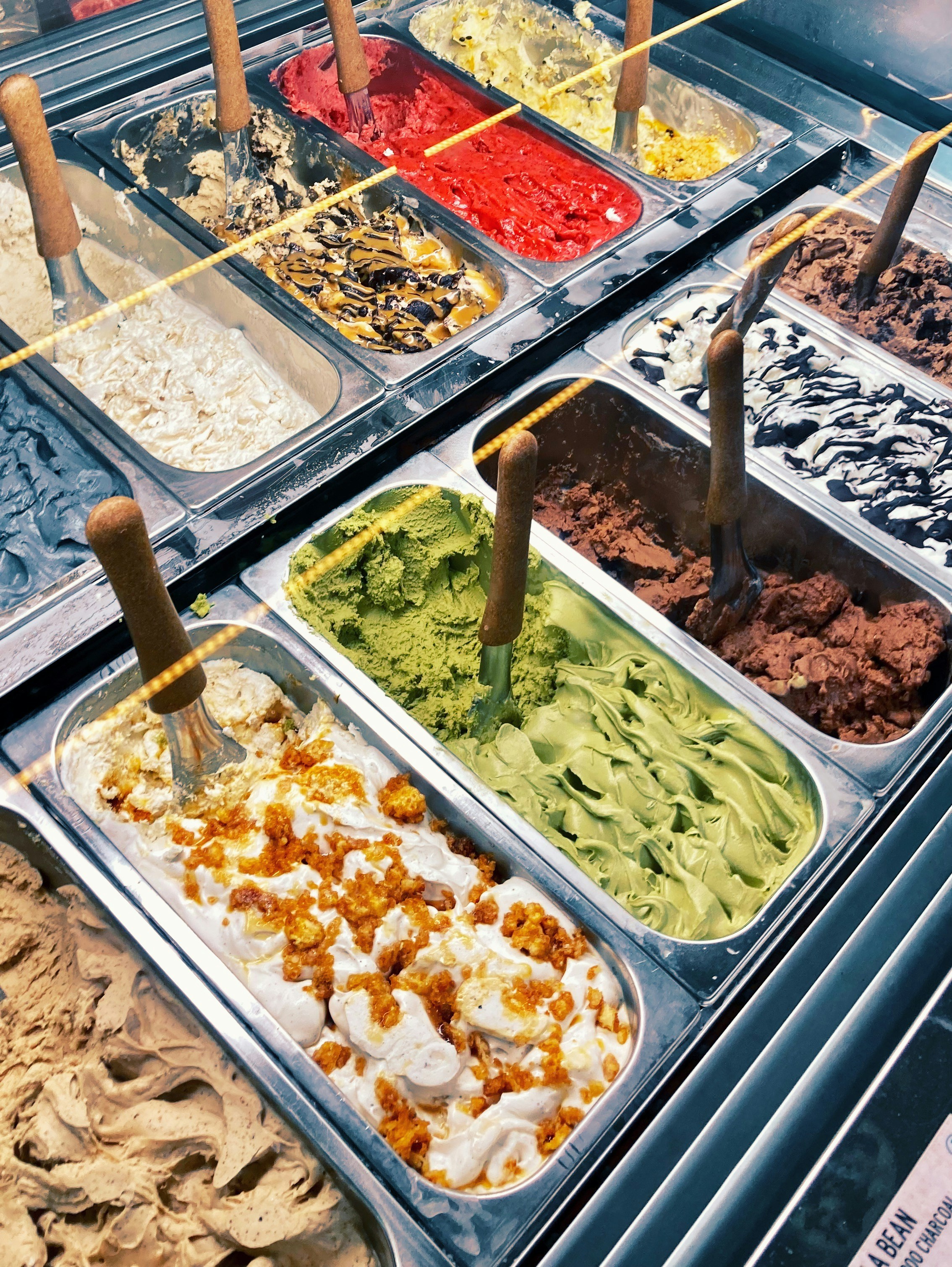 Display of assorted gelato flavours in metal tubs with scoops, including pistachio and chocolate.