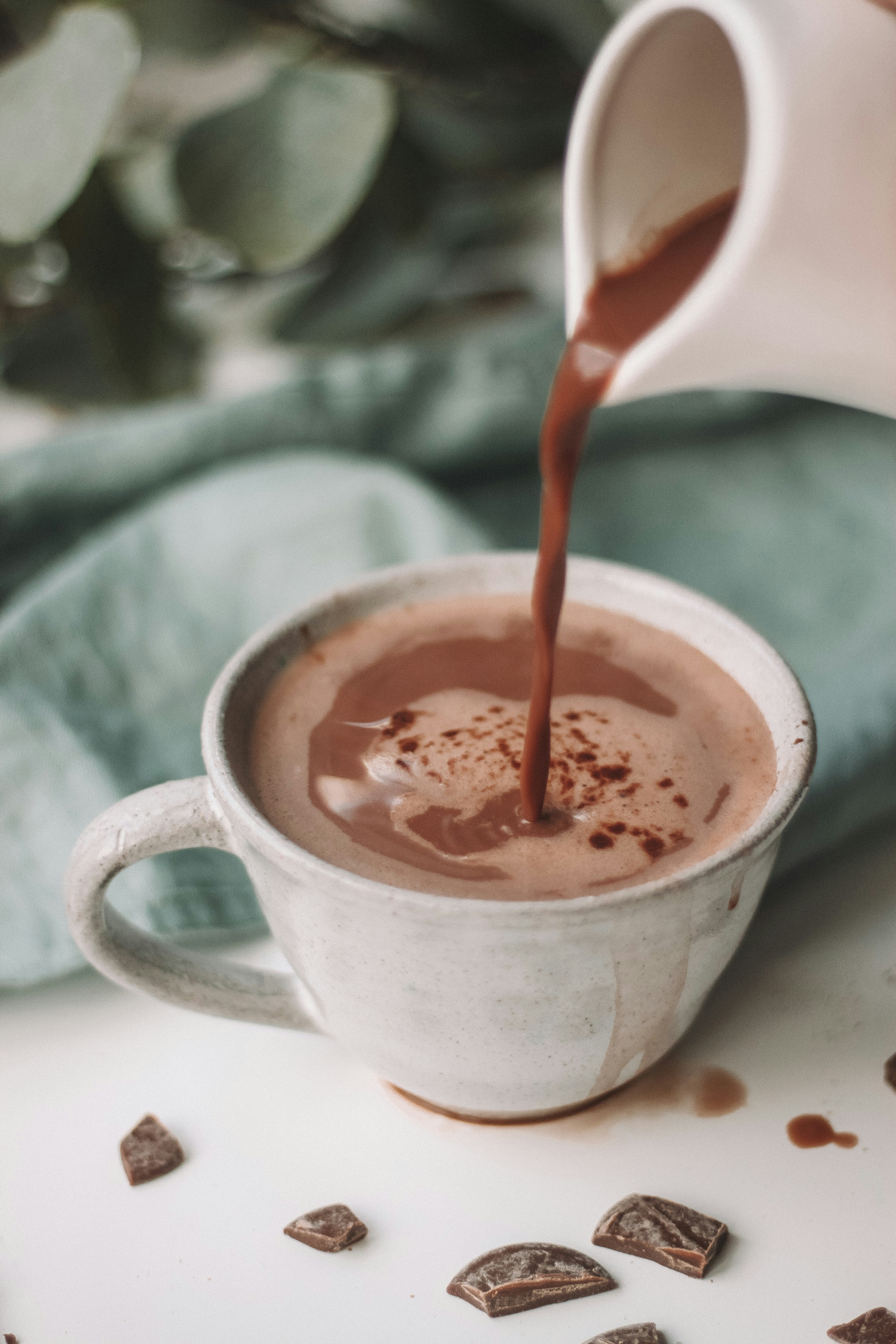 Hot chocolate being poured into a white mug on a light surface.