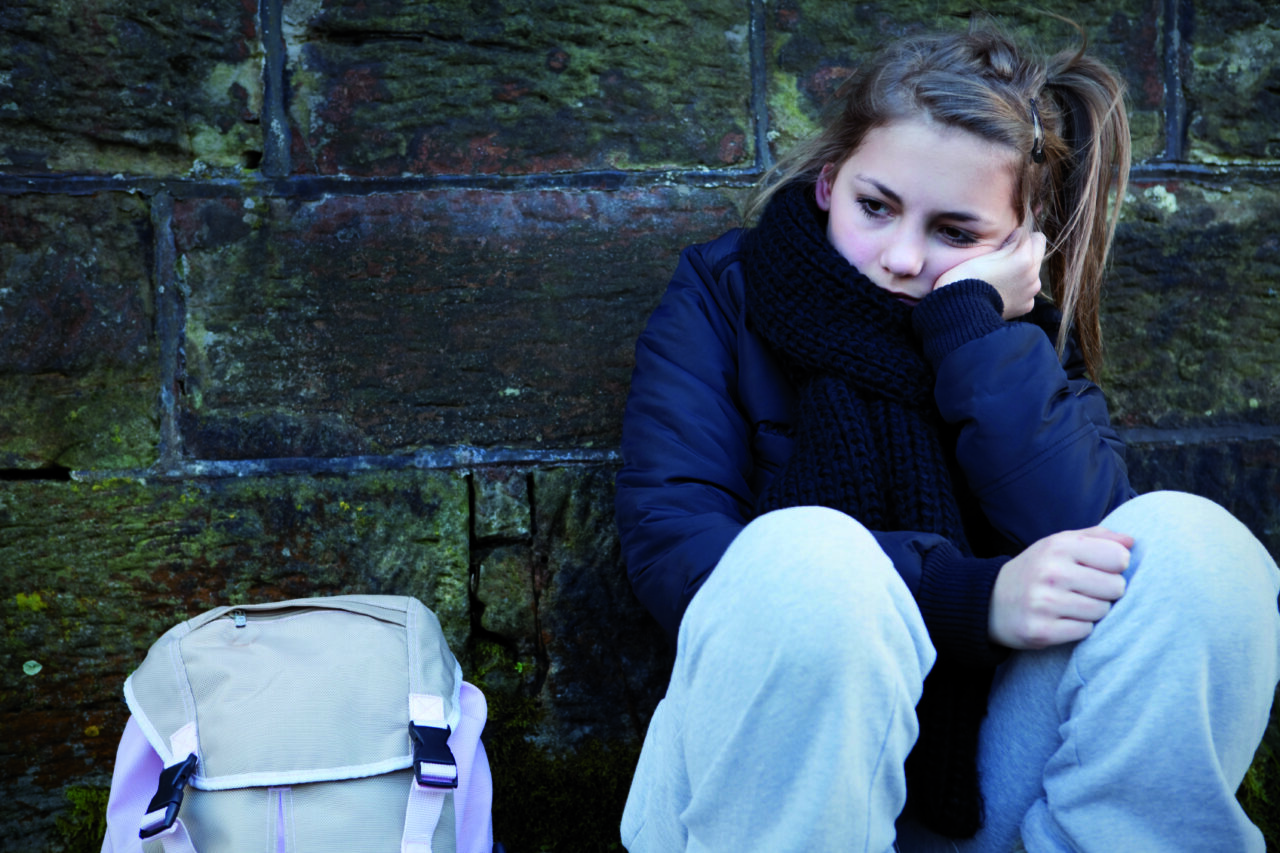 Young person sitting outdoors against a stone wall wearing a coat and scarf, looking cold and upset.