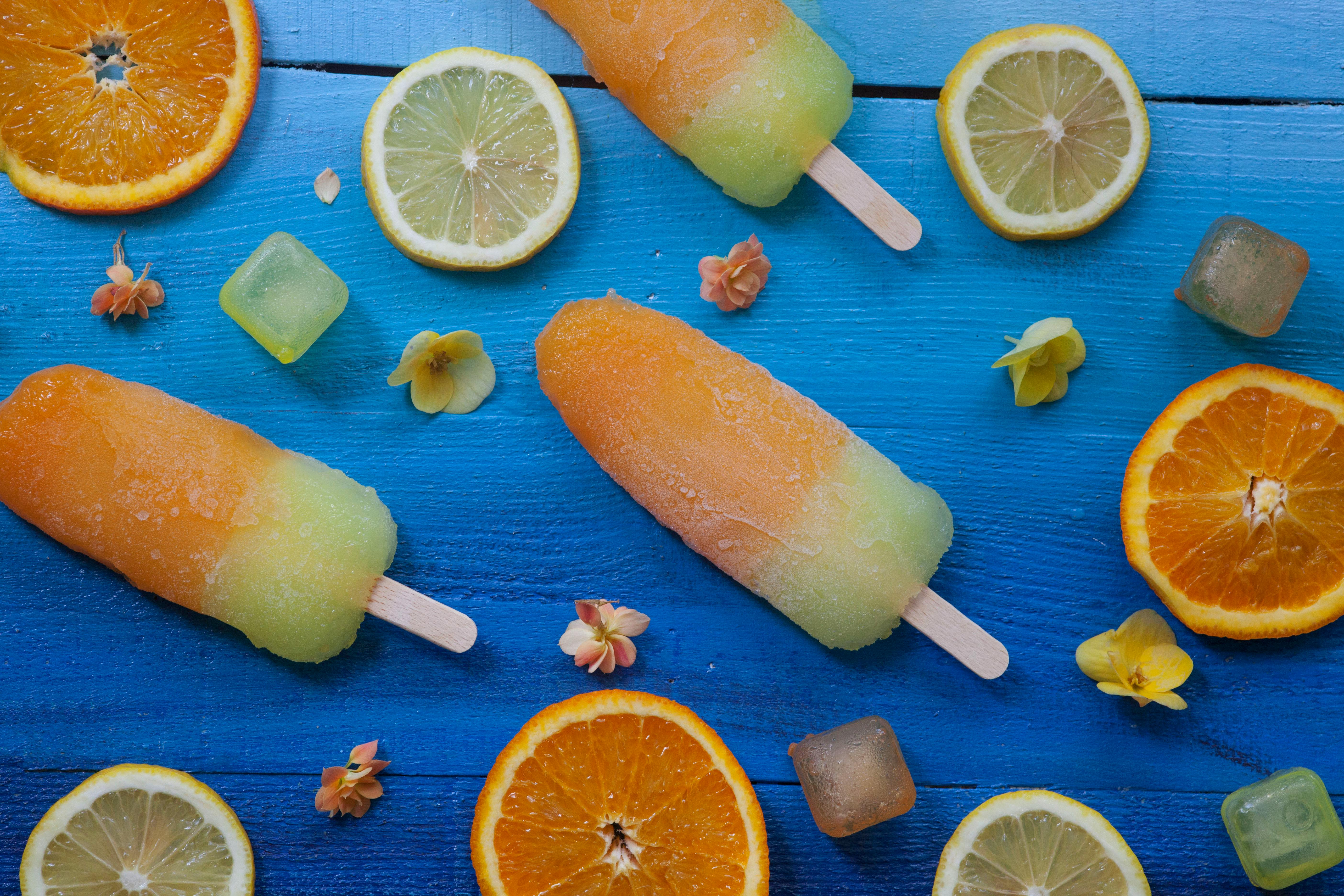 Orange and green ice lollies on a blue surface surrounded by slices of fruit and flowers.