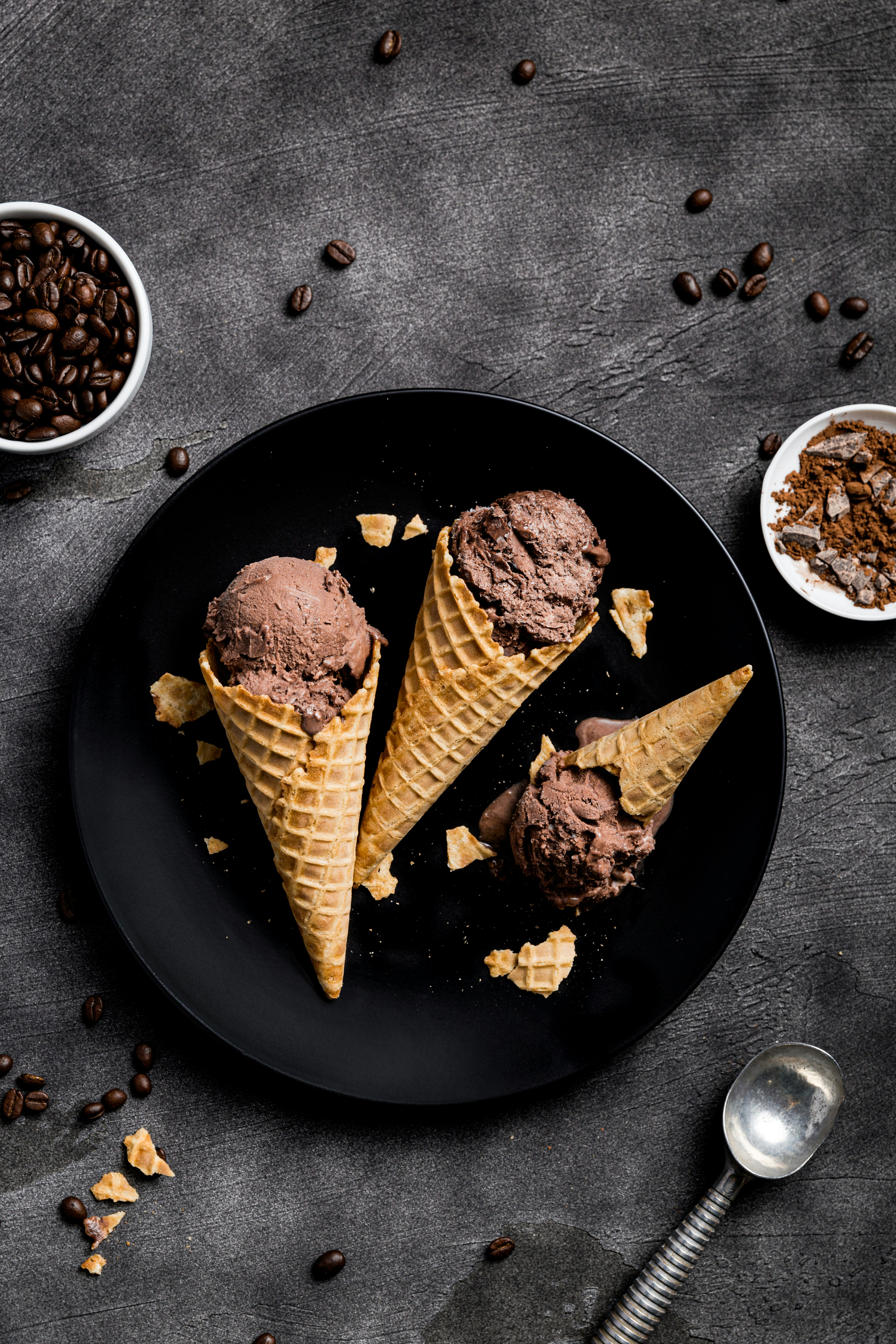 Three cones of chocolate ice cream on a black plate surrounded by pieces of cone.