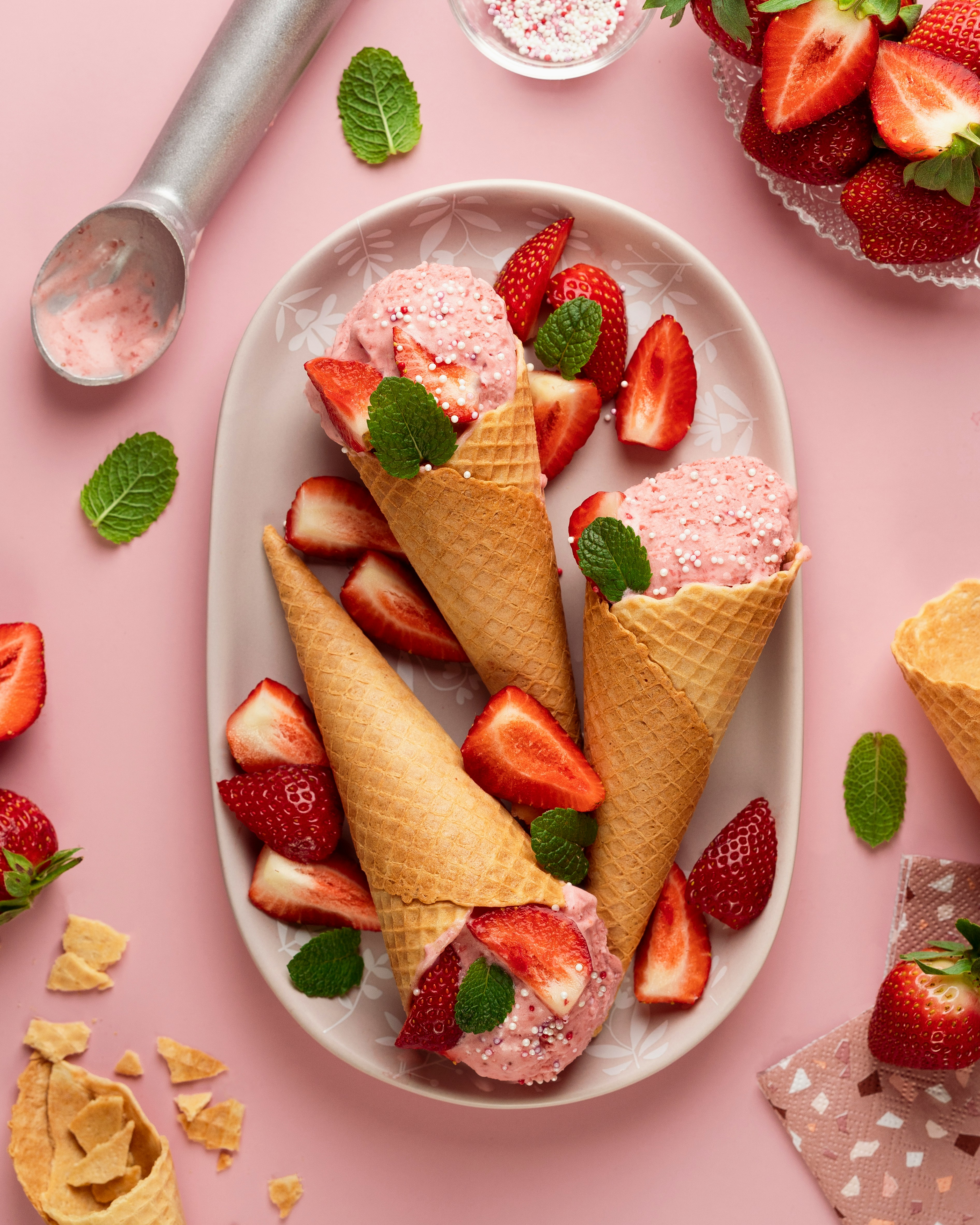 Three strawberry ice cream cones on a plate topped with sprinkles, strawberries, and mint leaves.