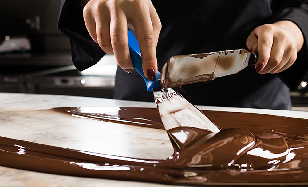 A woman wearing black tempers milk chocolate by hand on a silver surface using two metal scrapers.