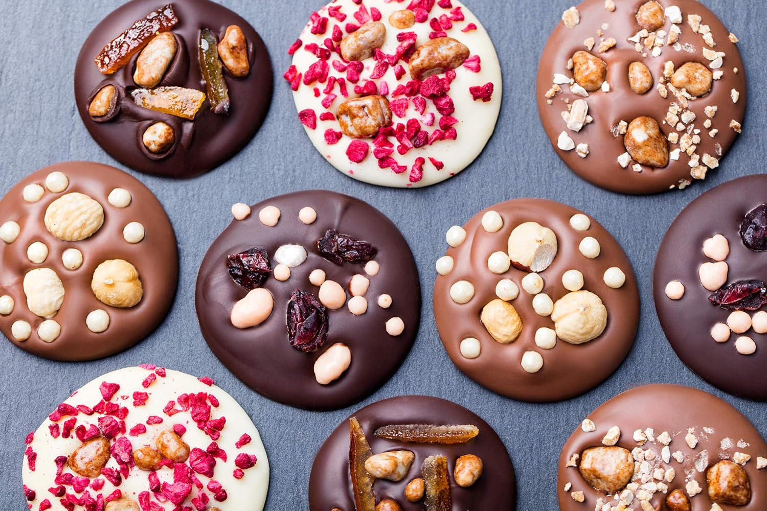 Rounds of deposited milk, dark and white chocolate with assorted toppings on a dark surface.